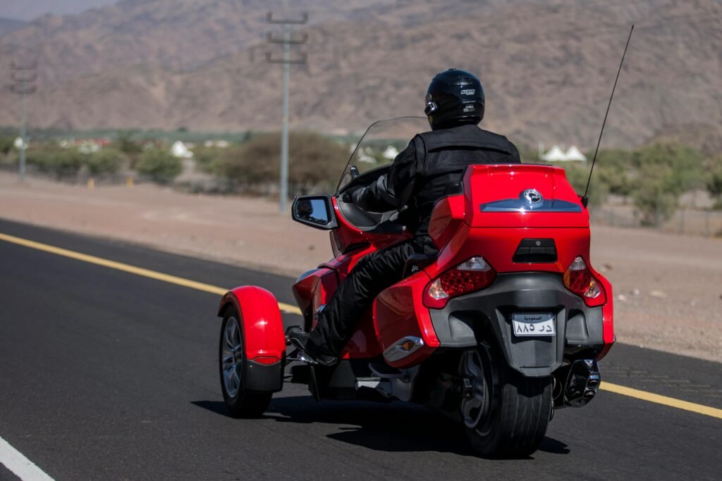 A motorcyclist rides a red trike on a desert highway, enjoying a sunny day with mountainous backdrop.